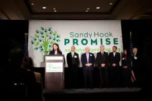 A speaker stands at a podium with several people behind, in front of a "Sandy Hook Promise" banner.