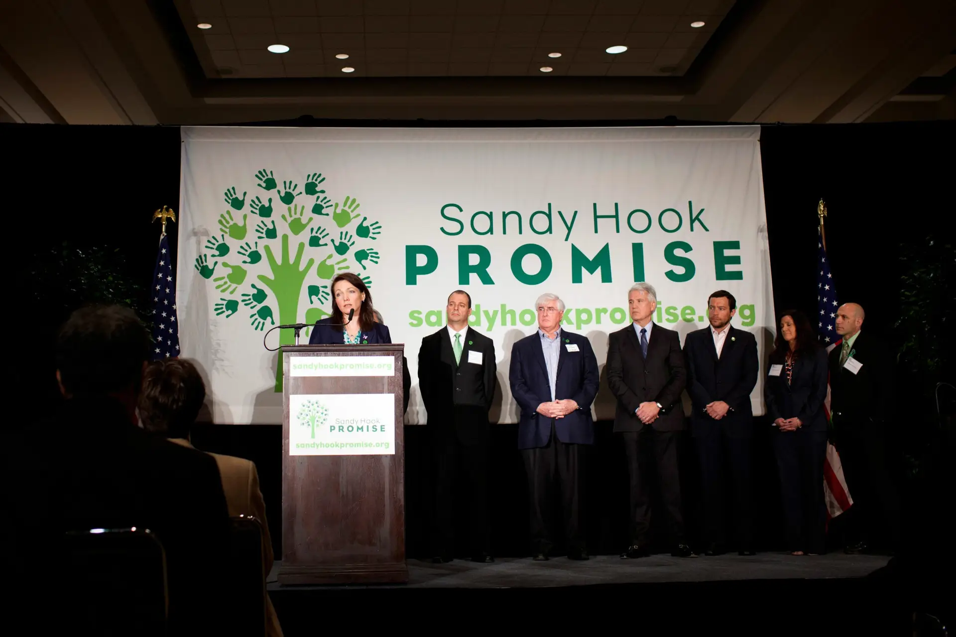 A speaker stands at a podium with several people behind, in front of a "Sandy Hook Promise" banner.