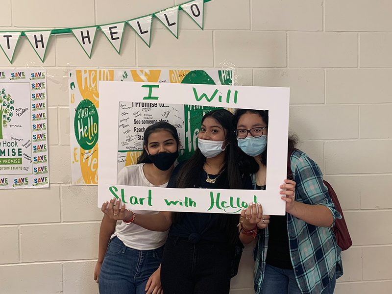 The image shows three students within a photo frame. The frames says “I will Start With Hello!