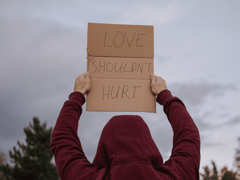 man in a hoodie holding up a sign with the words "love shouldn't hurt"