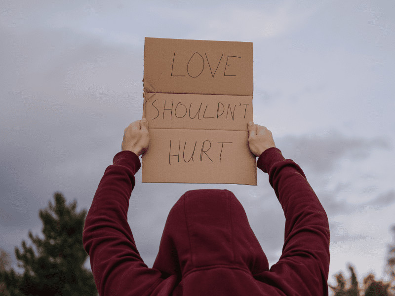 Man in a hoodie holding up a sign with the words "love shouldn't hurt"