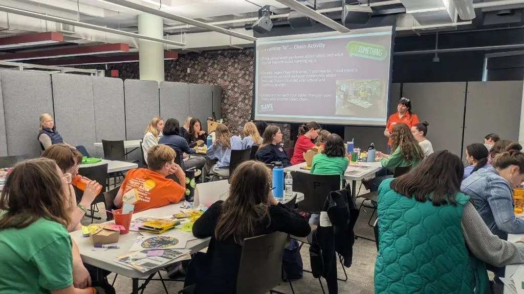 A group of young women is engaged in an interactive workshop for a SAVE Promise Club youth leadership event. A presenter in an orange shirt is leading an activity called “I Promise To…” which is displayed on a screen. Participants are seated at tables filled with arts and crafts materials, papers, and snacks. The environment is collaborative, with students actively writing and discussing.