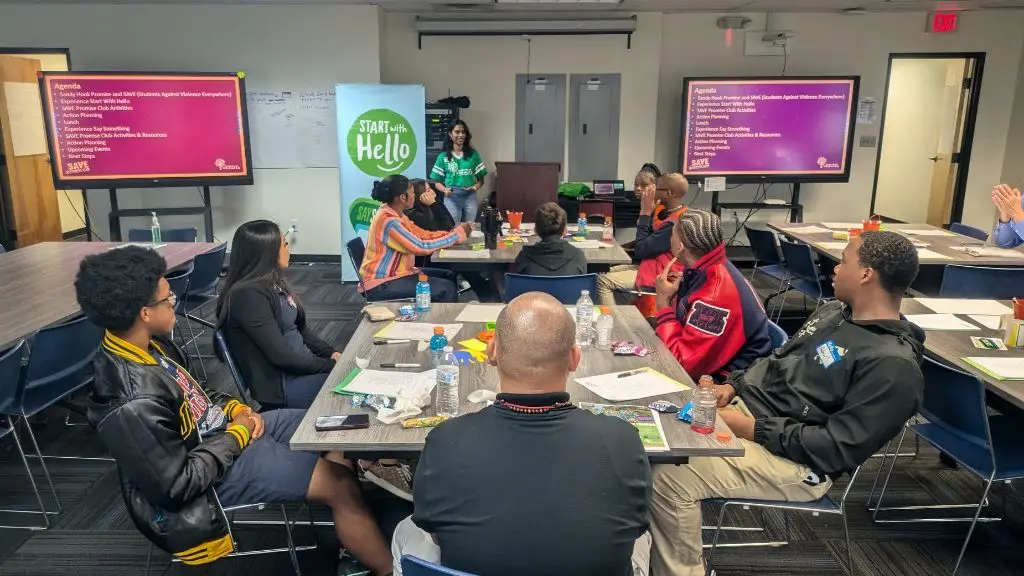 A diverse group of young people and adults are seated at tables in a conference-style room for a SAVE Promise Club youth leadership event, attentively listening to a presenter at the front. The presenter, a woman in a green shirt, stands behind a podium with a "Start With Hello" banner nearby. Large screens display an agenda related to violence prevention and youth leadership. The attendees have papers, snacks, and water bottles on their tables.