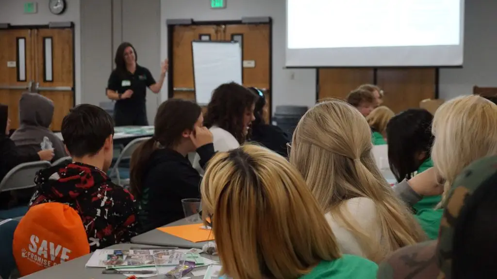 A group of young students is engaged in a SAVE Promise Club youth leadership event. A presenter in an black polo shirt stands at the front, leading an activity. The environment is collaborative, with students actively writing and discussing.